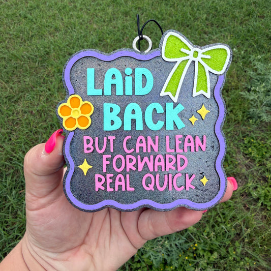 A hand holds a decorative sign outdoors with grass in the background. The sign has a dark gray background with a purple border, turquoise and pink text reading 'Laid Back But Can Lean Forward Real Quick,' a green bow graphic, and a yellow flower graphic.
