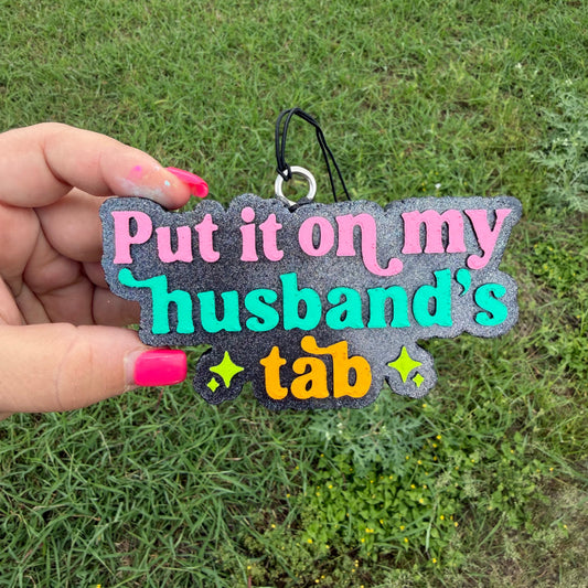 A hand with pink nail polish holds a glittery black keychain outdoors on a green grass background. The keychain features the text 'Put it on my husband's tab' in pink, teal, and orange lettering with yellow star accents.